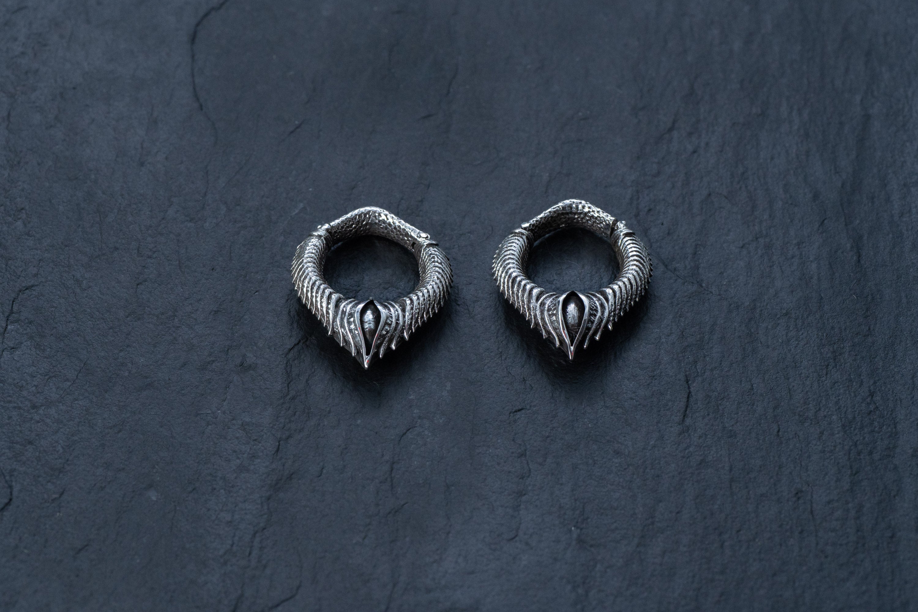 A pair of intricately designed MARVDASHT Small Biomechanical Clicker Ear Weights in 925 Silver by Kaya Movement, featuring curved, snake-like bodies and scale patterns, are displayed on a dark, textured background. These unisex 2 gauge earrings exude subtle sci-fi vibes.